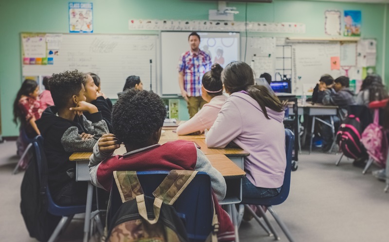 Une salle de classe pleine d’élèves, représentant une gamme de races et de genres, avec un enseignant à l’avant.